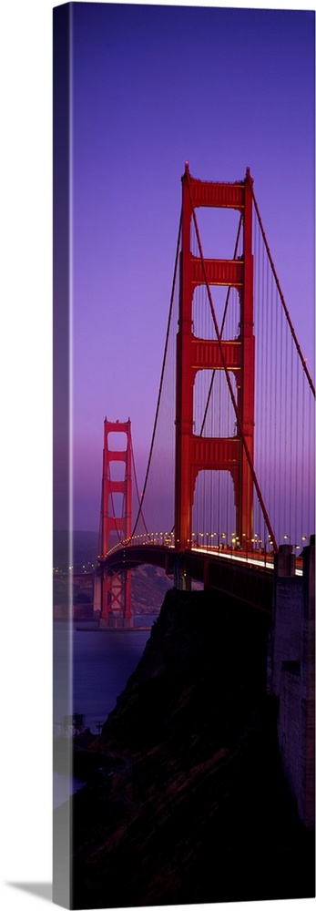 Oversized, vertical photograph taken at an angle of the Golden Gate Bridge at dusk, in San Francisco, California.