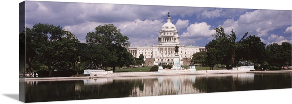 Panoramic photograph of municipal building at water's edge surrounded by trees under a cloudy sky.