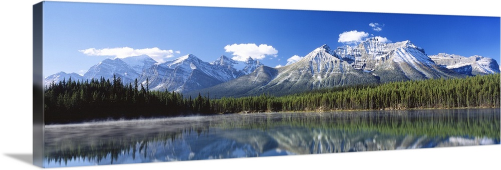 Large panoramic canvas of big snowy mountains with a dense forest beneath it reflected in the water.