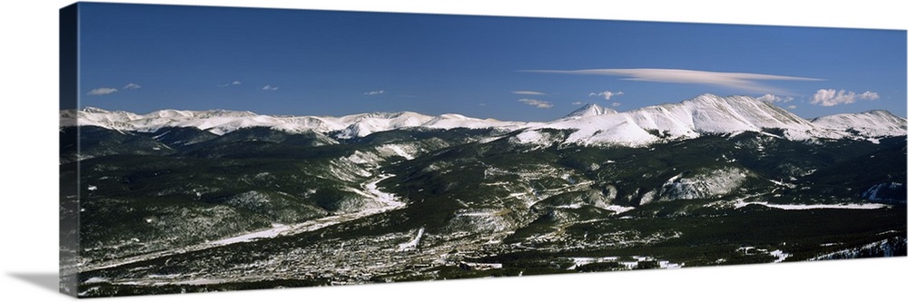 This wide angle photograph is an aerial shot of a snow covered mountain range with vast land and a town in front of them.