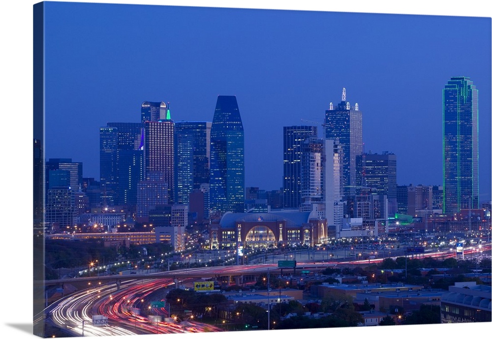 Big photograph of downtown Dallas, Texas (TX) at night with a busy road in the foreground in front of towering skyscrapers.