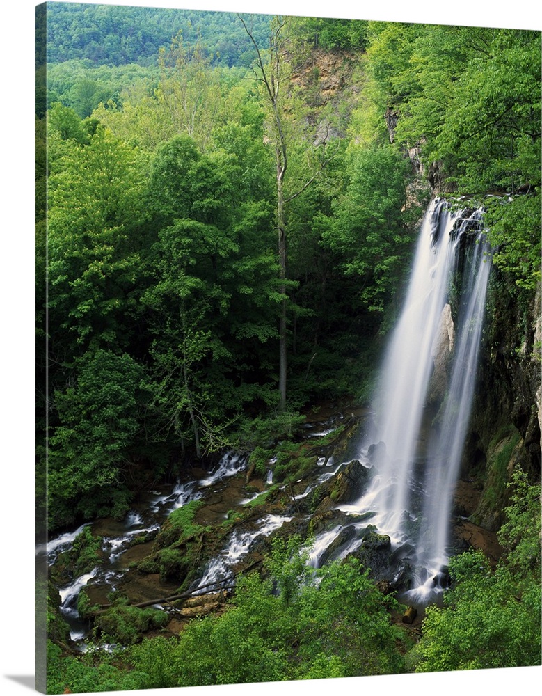A large waterfall is photographed from above as it falls onto rocks and is surrounded by thick forest.