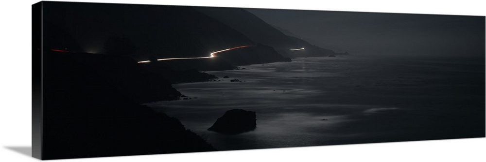 Panoramic photograph of road near coastline lit up with moving headlights at night.