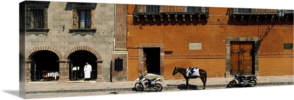 This panoramic photograph shows two motorcycles sitting on a stone street with a horse standing in between them. Parts of ...
