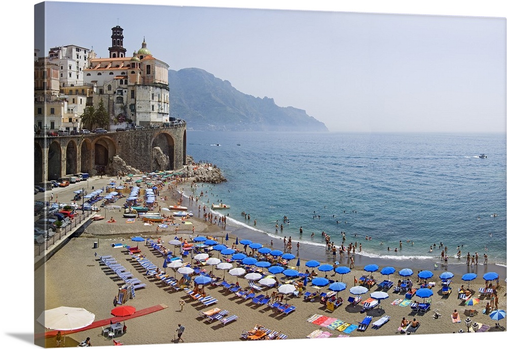 Summer scene on the beach with lots of parasols and swimmers, and historic buildings on the edge of the ocean.