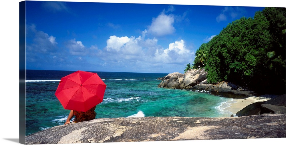 A woman holding a red umbrella sits on a large rock and looks out over the ocean with the coast just to her right.
