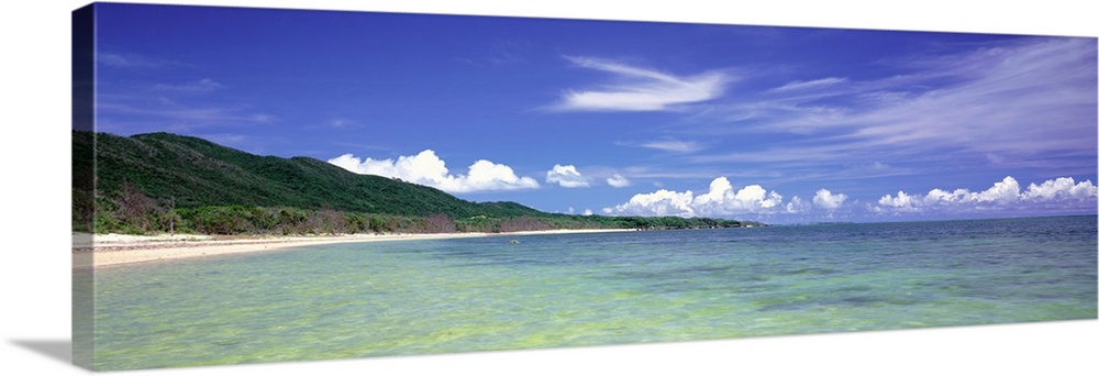 Panoramic photograph of ocean with shoreline and grass covered mountains in the distance under a cloudy sky.