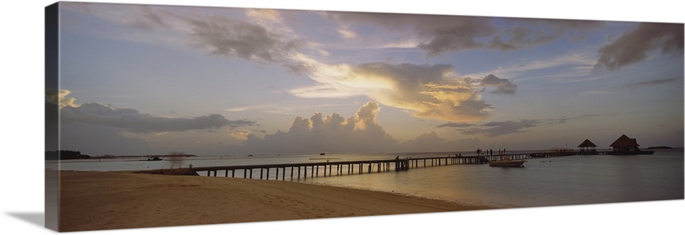 Panoramic photograph of pier stretching into ocean from the beach under a cloudy sky at dusk.