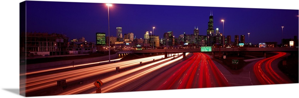 Panoramic photograph of highway filled with light trails with lit up city skyline in the distance at night.