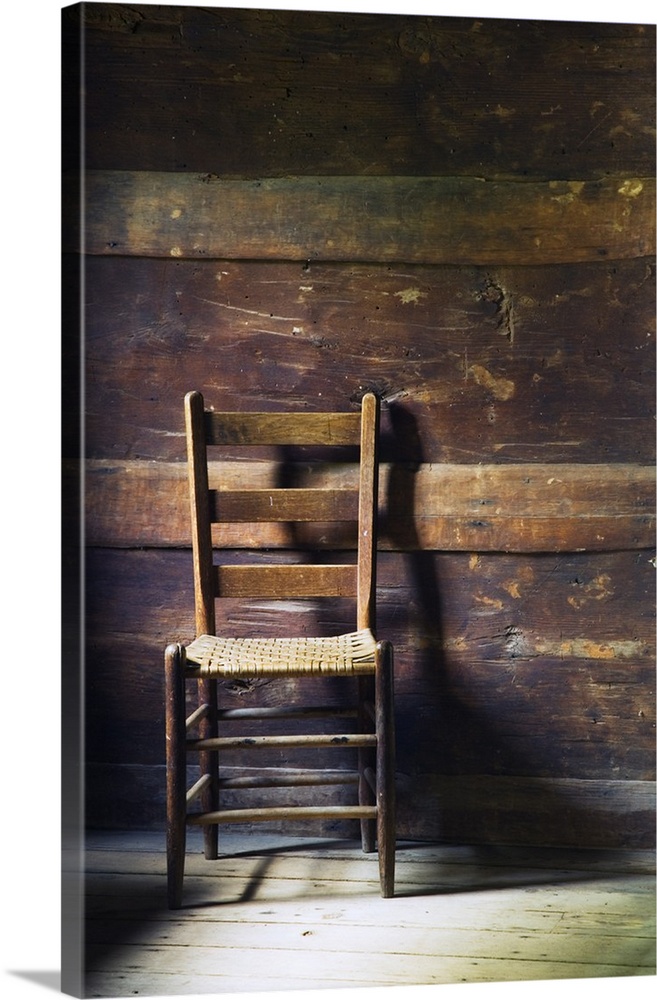 Portrait photo of an empty ladderback wooden chair sitting in a beat up wood slat wall room with sanded down floors.