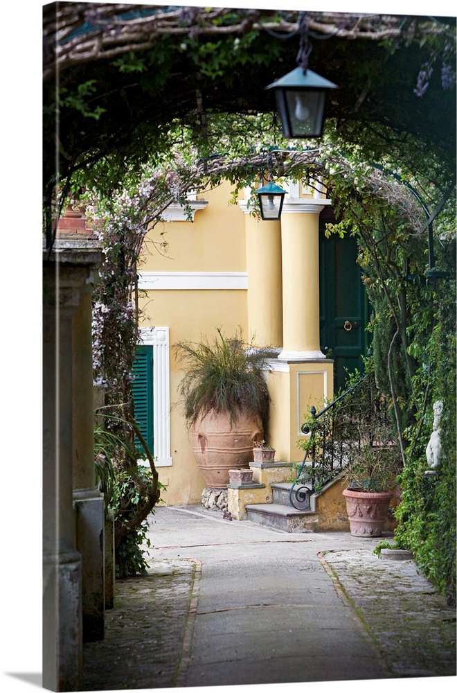 A vertical photograph or a walkway under vine covered arches in a scenic Italian city.