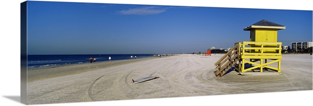 A panoramic photograph of a mostly empty beach, there is a small shelter and abandoned surfboard in the foreground of the ...