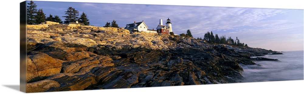 A large panoramic photograph taken from sea level and looking up at a lighthouse. Rocks take up most of the foreground wit...