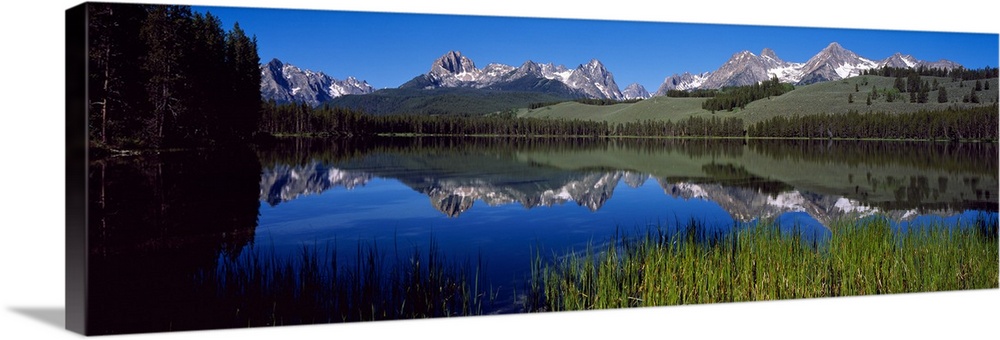 A mountainous view sits behind a hill and reflects in the water that the picture has been taken across from.