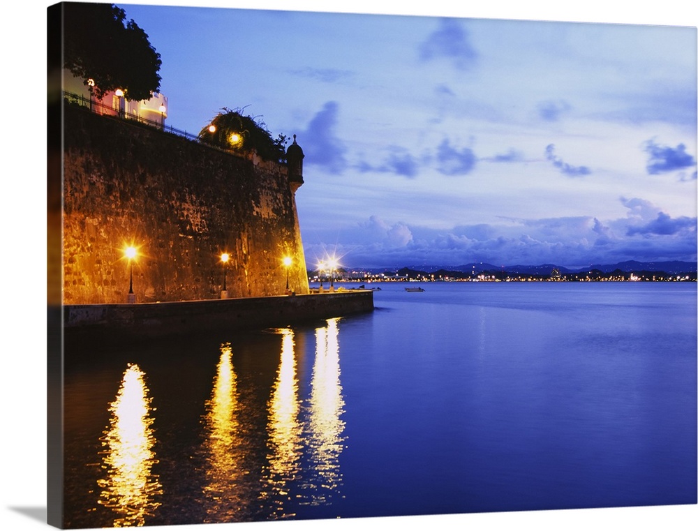 Photograph taken over a body of water with a coast lit up in the distance and to the left a large stone wall with a buildi...