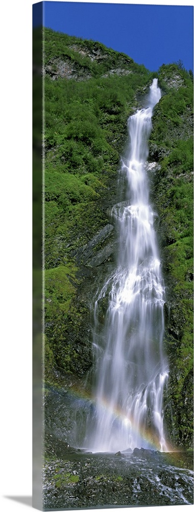 Vertical panoramic photograph of water cascading over rocks with small rainbow at the bottom.