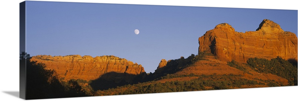 A panoramic photograph of the setting sun illuminating rock cliffs as the moon rises between them.