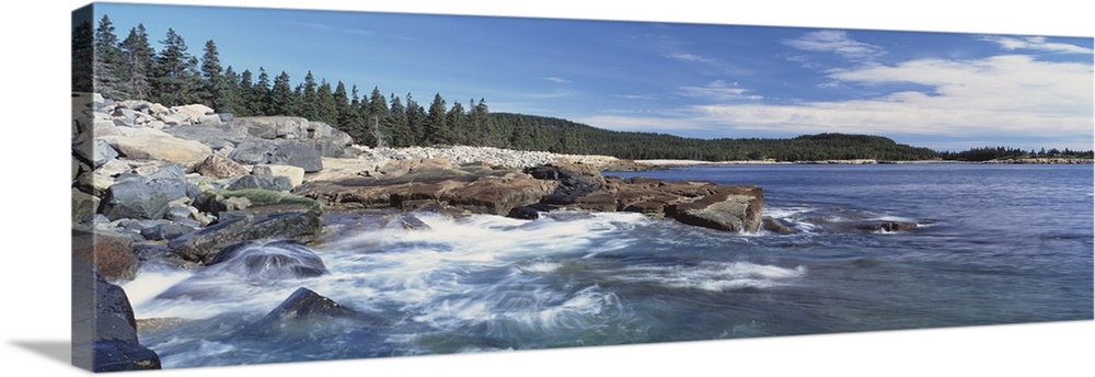 Panoramic photograph of rocky shoreline with surf.  There are trees in the distance under a cloudy sky.
