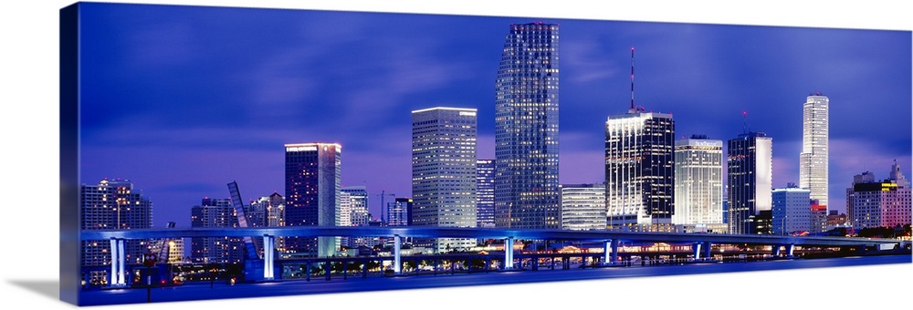 Big panoramic canvas photo of a cityscape on the water with a long bridge passing in front from right to left at dusk.