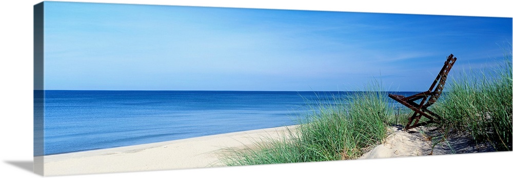 This wall art is a panoramic landscape photograph of a sandy beach with a chair in the dunes overlooking the lake.