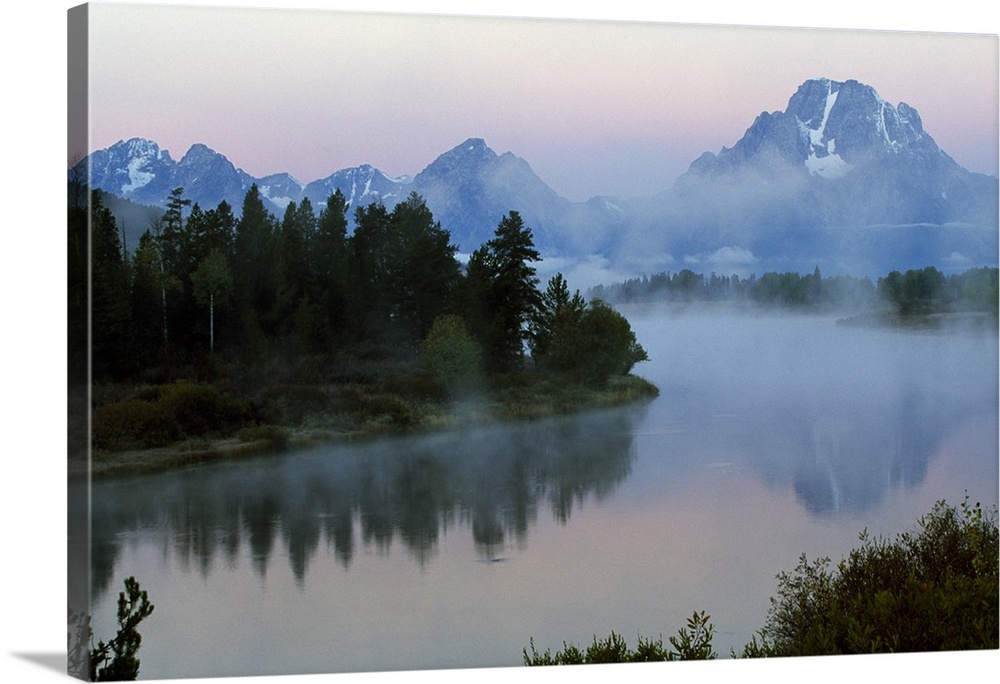 A large photograph showing forest and foilage surrounding water with mountains in the background and mist floating above.