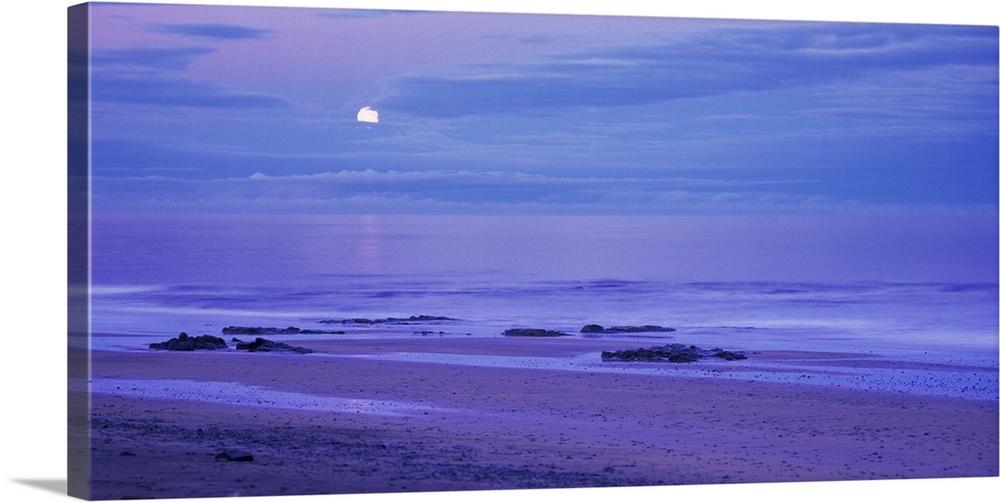 The moon is seen slightly behind a cluster of clouds that hang over the ocean water and photographed in a wide angle view.