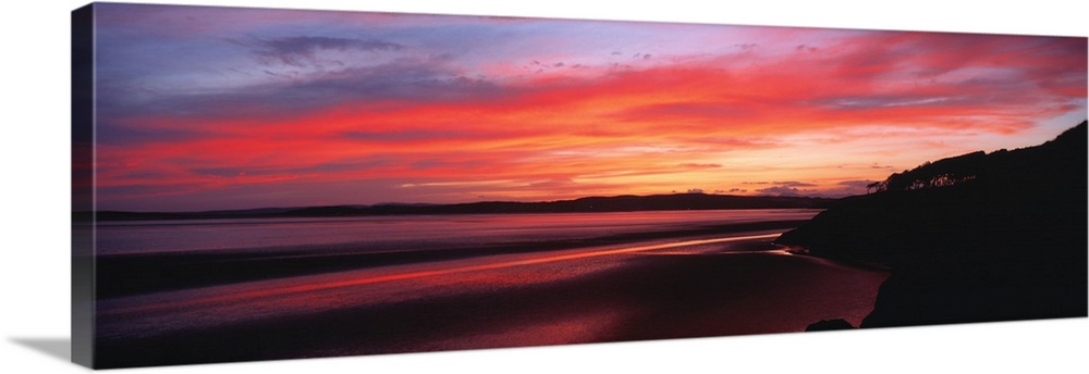 A large panoramic photograph taken of a sunset over a bay in the UK. A hill top is silhouetted to the very right of the pr...