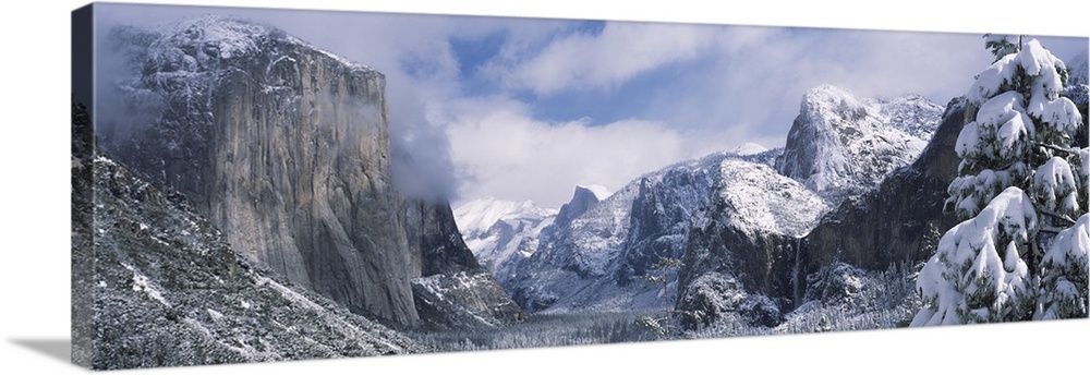 This panoramic photograph is taken of immense mountains that reach up into the clouds and are covered with snow.