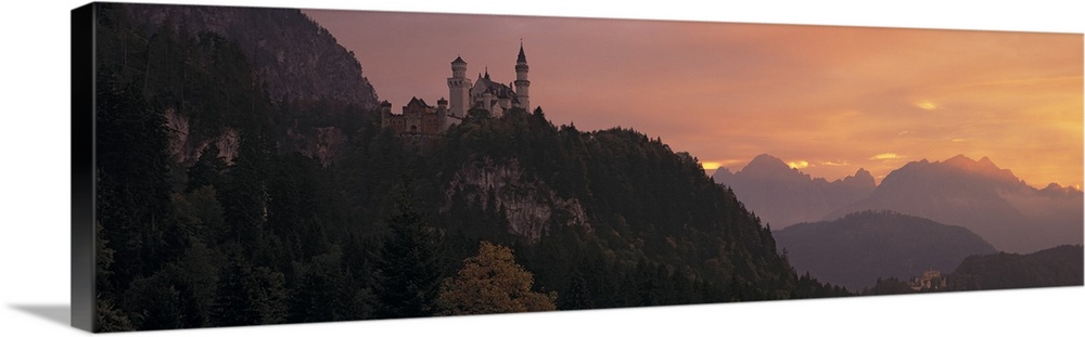 A large panoramic piece of a castle sitting in the mountains in Germany. The sky has warm tones as the sun sets out of view.