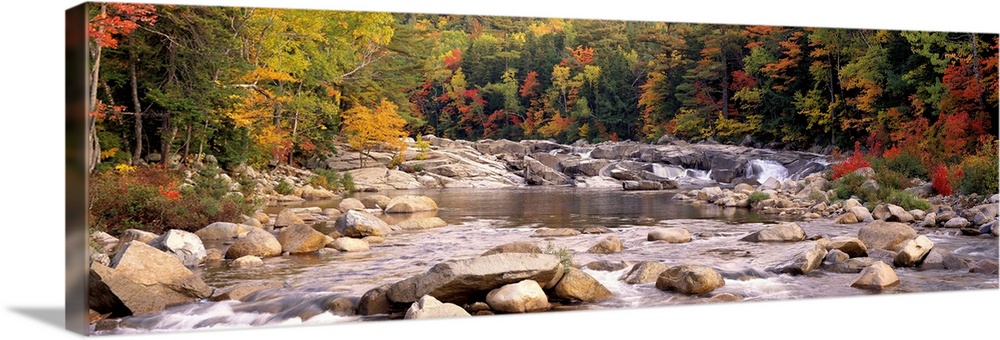 A relaxed panoramic landscape of large boulders in a New England river lined with trees with autumn colors.
