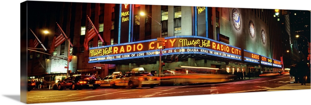 Panoramic view of Radio City Music Hall from the street corner at night, lit up by neon lights and with taxis passing by.