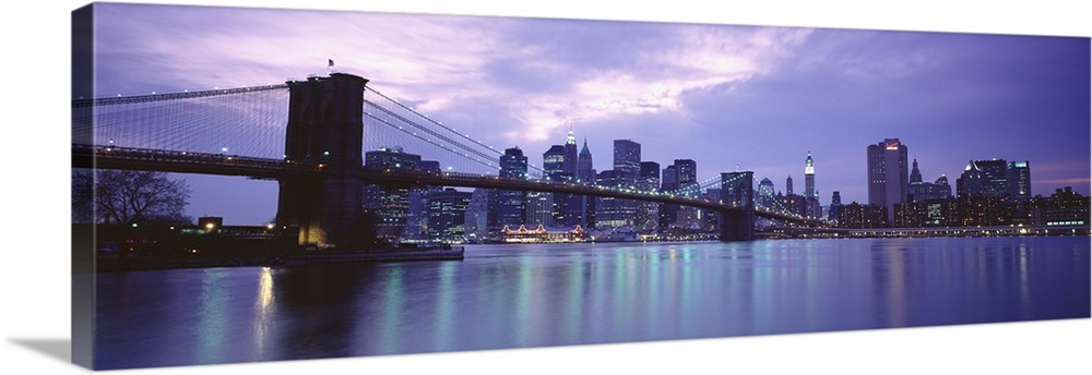 Large, panoramic photograph of the New York skyline, lit up at night, the Brooklyn Bridge in the foreground.