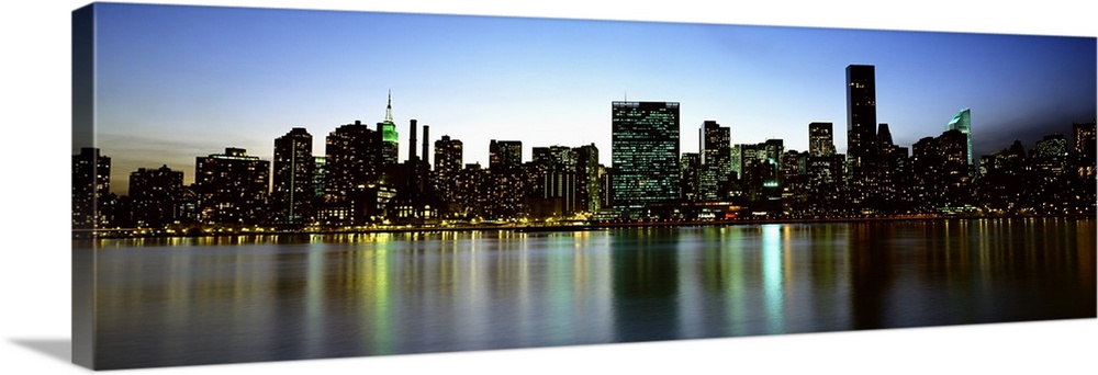 Panoramic photograph of the New York City skyline, lit at dusk and reflecting in the waters in the foreground.
