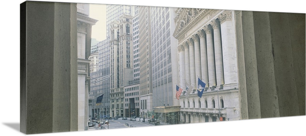 View between two concrete columns looking down Wall Street in New York City, with the faoade and columns of the Stock Exch...