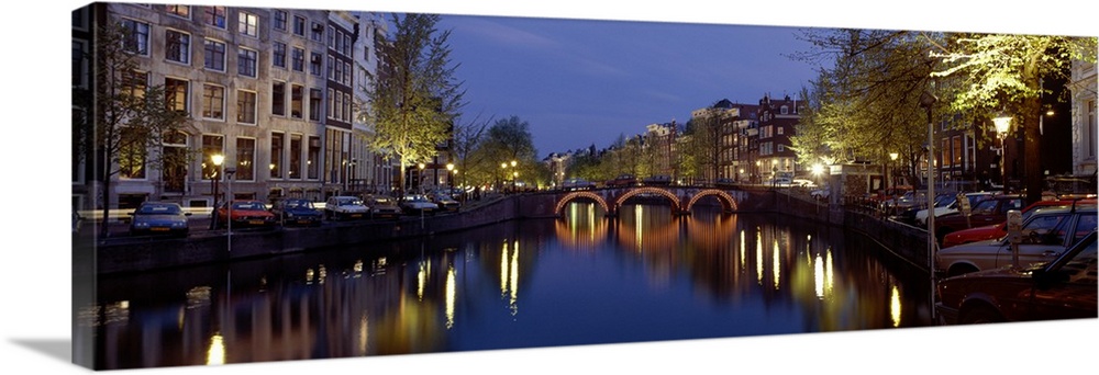 A wide angle view of a canal in Amsterdam with buildings and street lights illuminated on either side and reflecting in th...