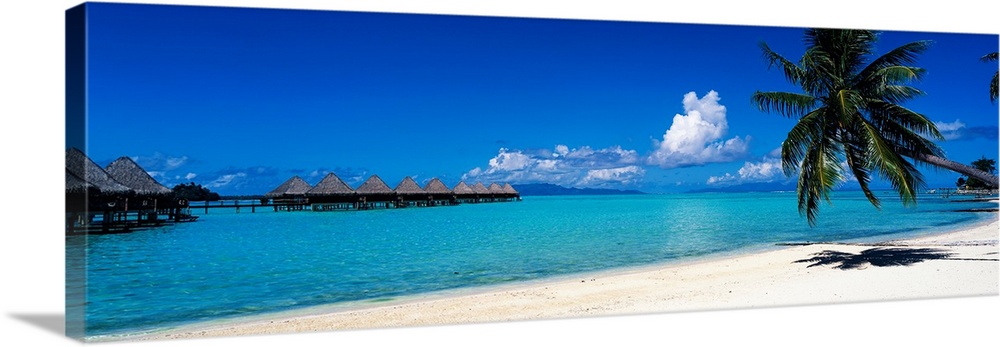 A panoramic photograph of a tropical beach with palm trees and thatch huts over the water.