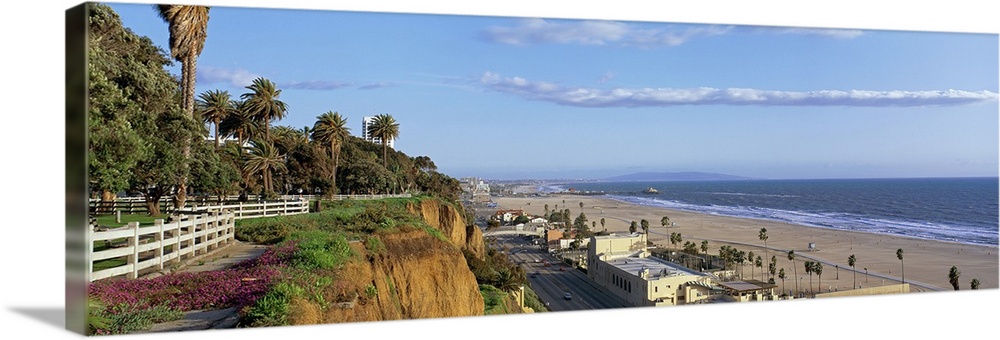Panoramic photograph of cliff overlooking shoreline filled with buildings and palm trees under a cloudy sky.