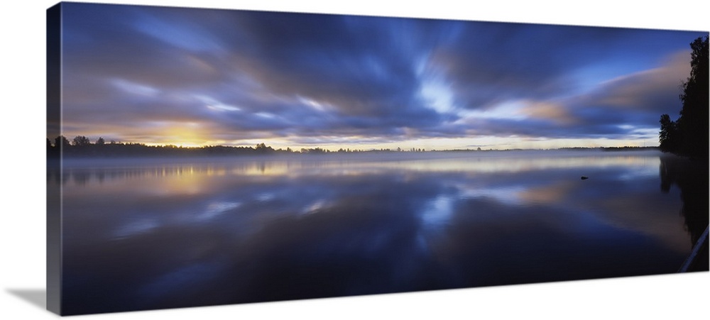 Panoramic photograph of huge body of water with trees in the distance at sunset.  The sky is cloudy and the clouds are ref...