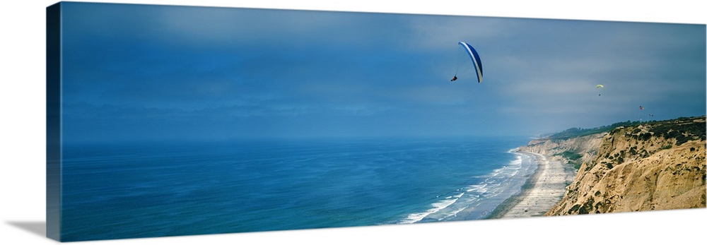 Horizontal photograph on a giant canvas of several paragliders floating over bright blue waters along the coast of La Joll...