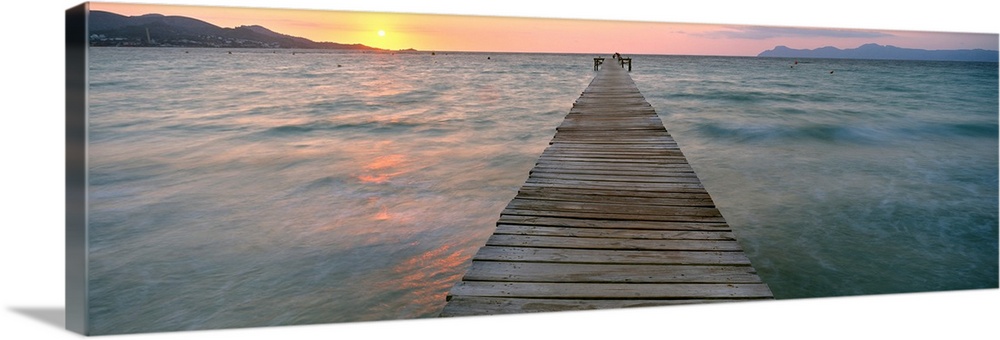 Panoramic photograph of wooden dock stretching into ocean at dusk with mountain silhouettes in the distance.