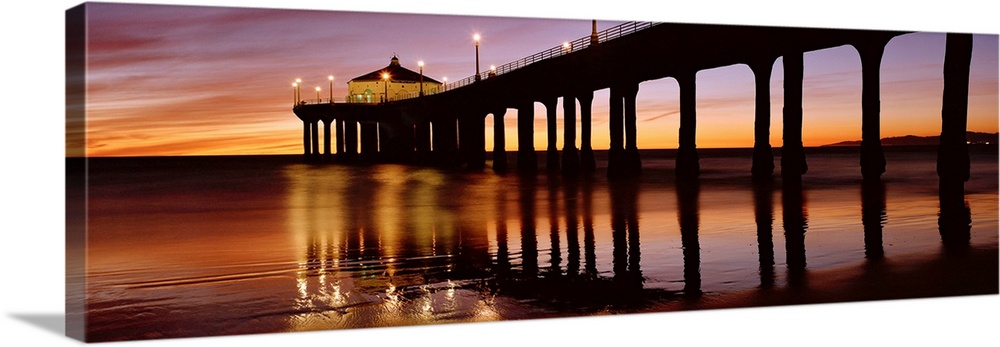 A panoramic photograph of the silhouette of a long pier over the ocean at sunset.