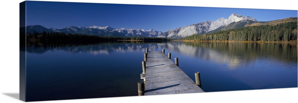 Panoramic photograph of tilted wooden dock stretching into the water.  There is a forest line and range of snow covered mo...