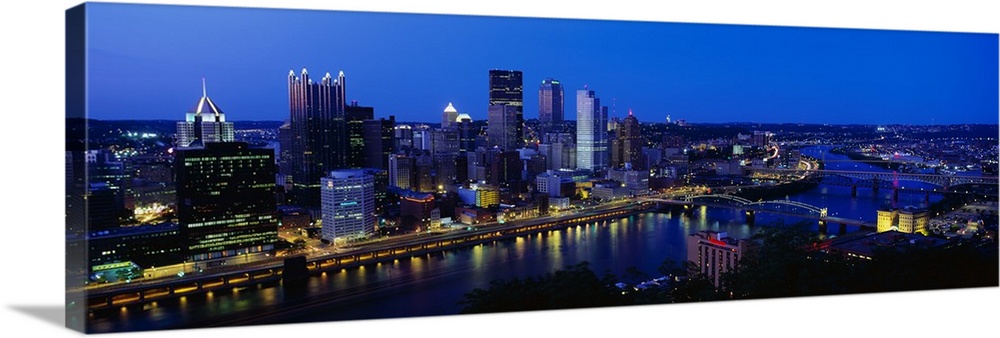 This huge panoramic photograph is of the Pittsburg skyline at night with the buildings lit up and a body of water in front...