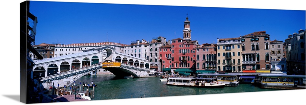 This panoramic view shows the Rialto bridge over the grand canal in Venice with buildings lining the water behind the bridge.