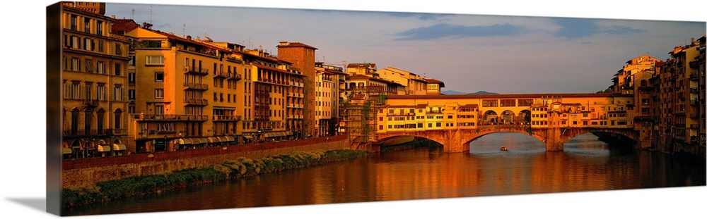Wide angle photograph of buildings alongside the Arno River, with a view of he Ponte Vecchio bridge, at sunset in Florence...