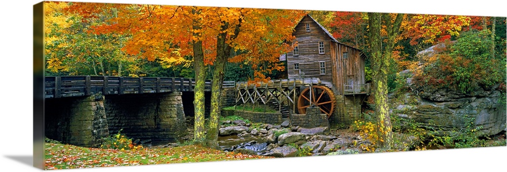 Panoramic picture taken of a mill through autumn colored trees with a bridge just to the left.
