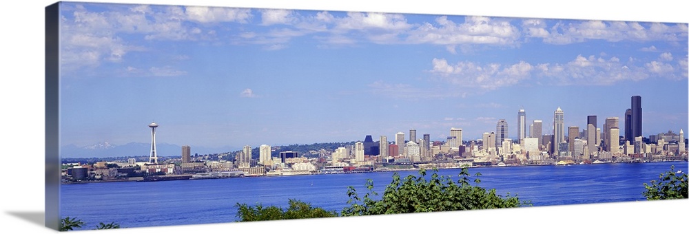A panoramic photograph is taken of the Seattle skyline across a large body of water with foliage at the bottom of the pict...