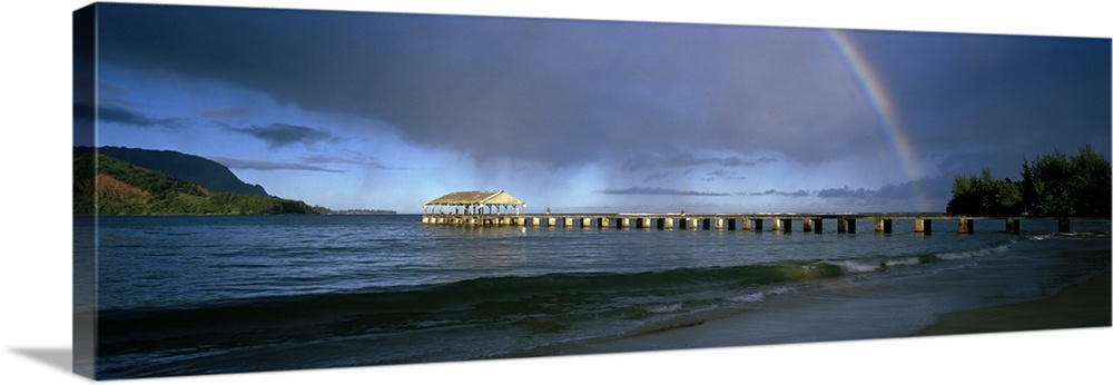 Panoramic photograph of long dock stretching into ocean with rainbow overhead.  There are mountains in the distance and da...