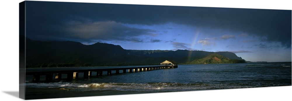 Picture taken of a long pier that reaches far out into the ocean with a faint rainbow shown near land that is in the dista...