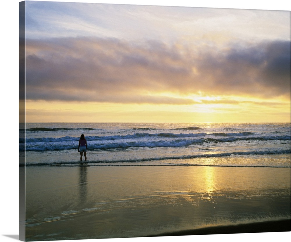 In this landscape photograph a lone figure stands in the waves on the shore watching the sunset over the horizon.